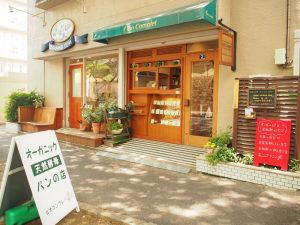 桜並木の木漏れ日の中で目を引く看板と、木とグリーンの相性がかわいらしいお店の外観