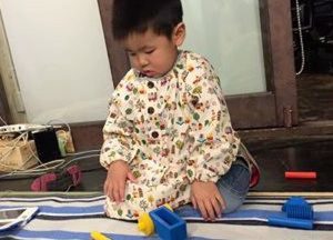 さっそくママ手作りのスモックに袖を通す男の子。「下の子が生まれ、家で落ち着いて作業することができない。子どもをみてくれているこの環境があったから作ることができた」とママ。直線縫いでできる袋物と違い、スモックは特に技術と時間がかかるグッズの一つだった12　写真キャプション（ずらりせいぞろい）
